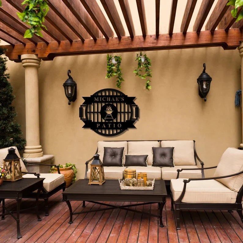Black steel grill master sign with sample name personalization displayed on tan patio wall above outdoor seating area with pergola