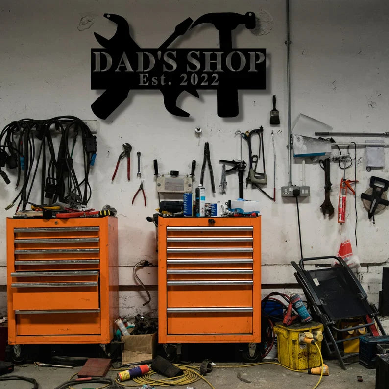 Custom metal workshop sign reading "DAD'S SHOP Est. 2022" mounted on a garage wall above orange tool chests and organized hand tools