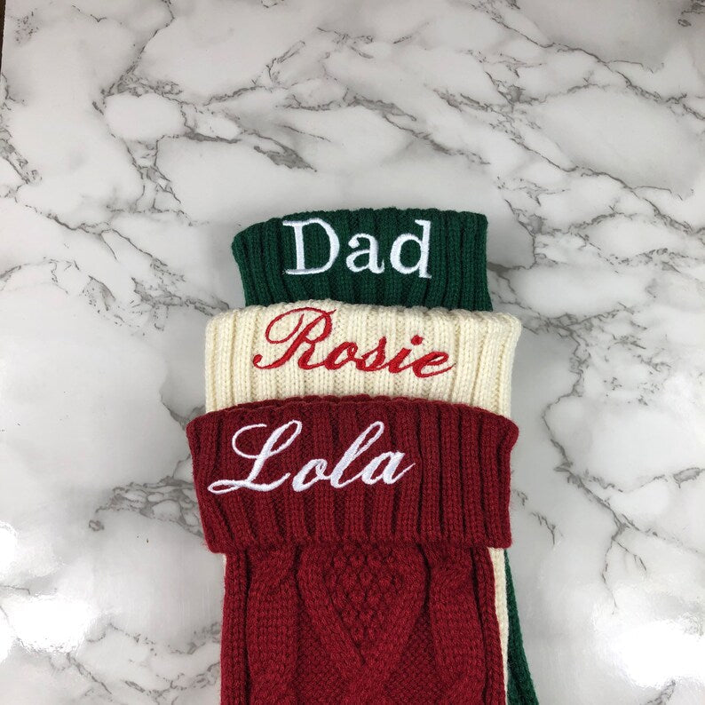 Three personalized knit stockings stacked flat, embroidered with sample names "Dad," "Rosie," and "Lola" in green, cream, and burgundy with ribbed cuff details