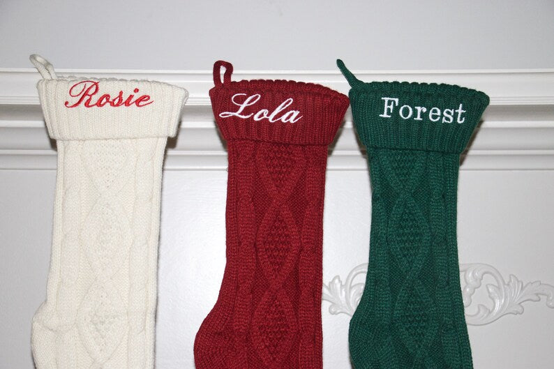 Three knit Christmas stockings in cream, burgundy, and forest green, each embroidered with a sample name: "Rosie," "Lola," and "Forest" respectively