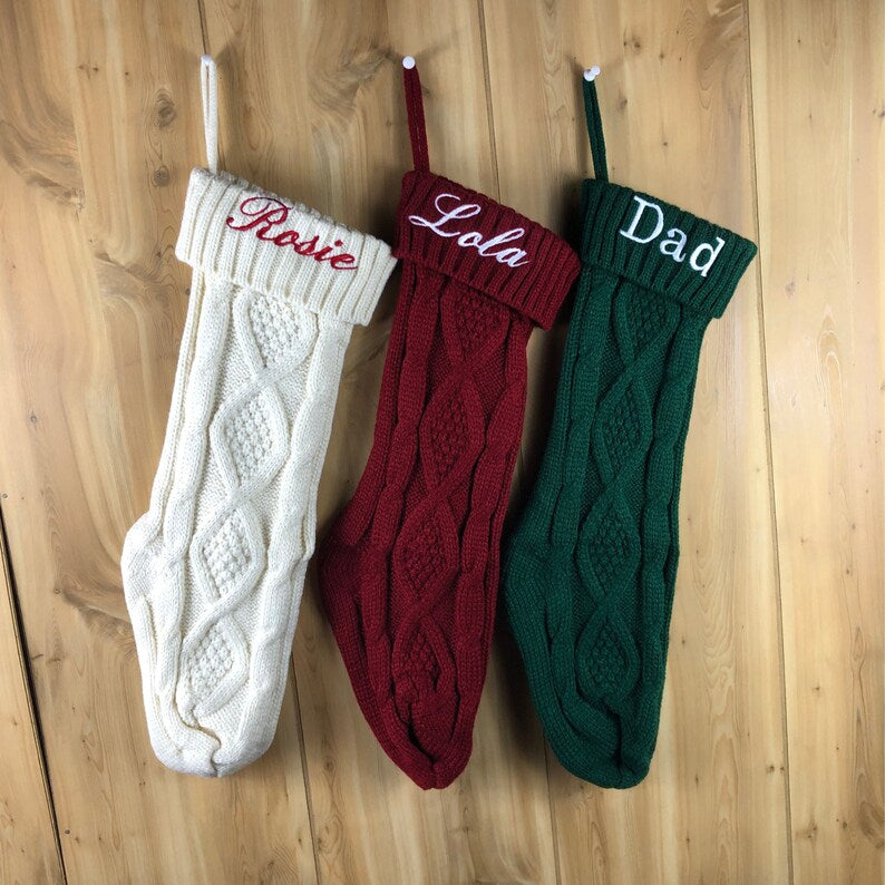 Three cable-knit Christmas stockings in white, burgundy, and forest green, each embroidered with a sample name—Rose, Lola, and Dad—hanging from white loops against a wooden background.
