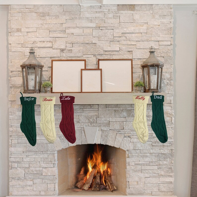 Fireplace mantel decorated with stone brick, displaying three empty picture frames, lanterns, potted plants, and six personalized knit stockings in green, cream, burgundy, and cream colors embroidered with sample names including Baylor, Rosie, Lola, Molly, and Dad.