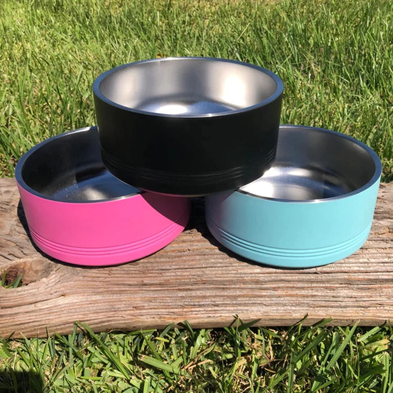 Three stainless steel dog bowls with colored exterior wraps—black, hot pink, and turquoise—displayed on weathered wood with grass background.
