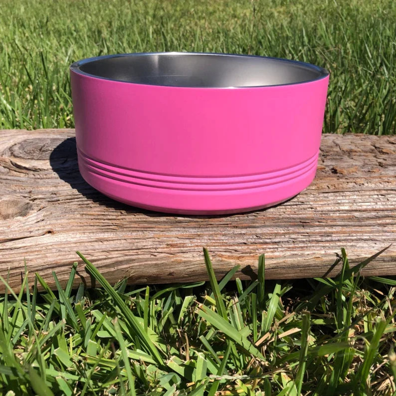 Bright pink stainless steel dog bowl with grooved detail band, displayed on weathered wood outdoors with grass background