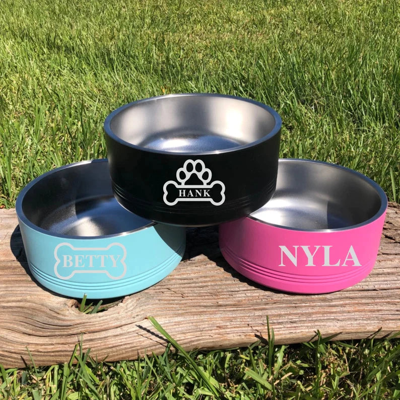 Three personalized stainless steel dog bowls in black, turquoise, and hot pink, engraved with sample names "Hank," "Betty," and "Nyla," displayed on wooden surface with grass background.