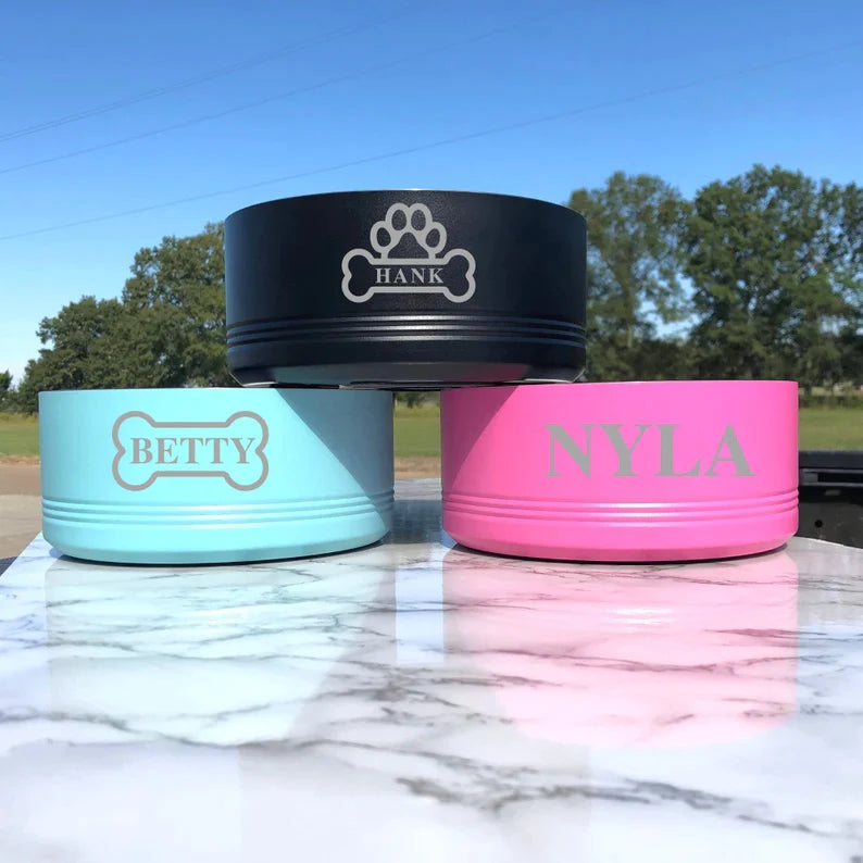 Three personalized dog bowls in black, light blue, and hot pink, each engraved with sample names "Hank," "Betty," and "Nyla" respectively, displayed on marble surface outdoors.