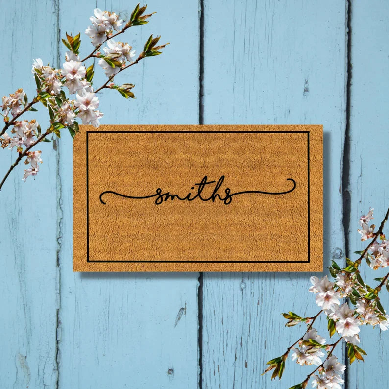 Tan coir doormat with black border and sample name "smiths" engraved in script, displayed on light blue wooden plank background with white flowering branches.