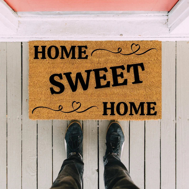 Natural coir doormat printed with "HOME SWEET HOME" text and decorative heart flourishes, shown placed on white wooden porch with person wearing black boots standing on it.