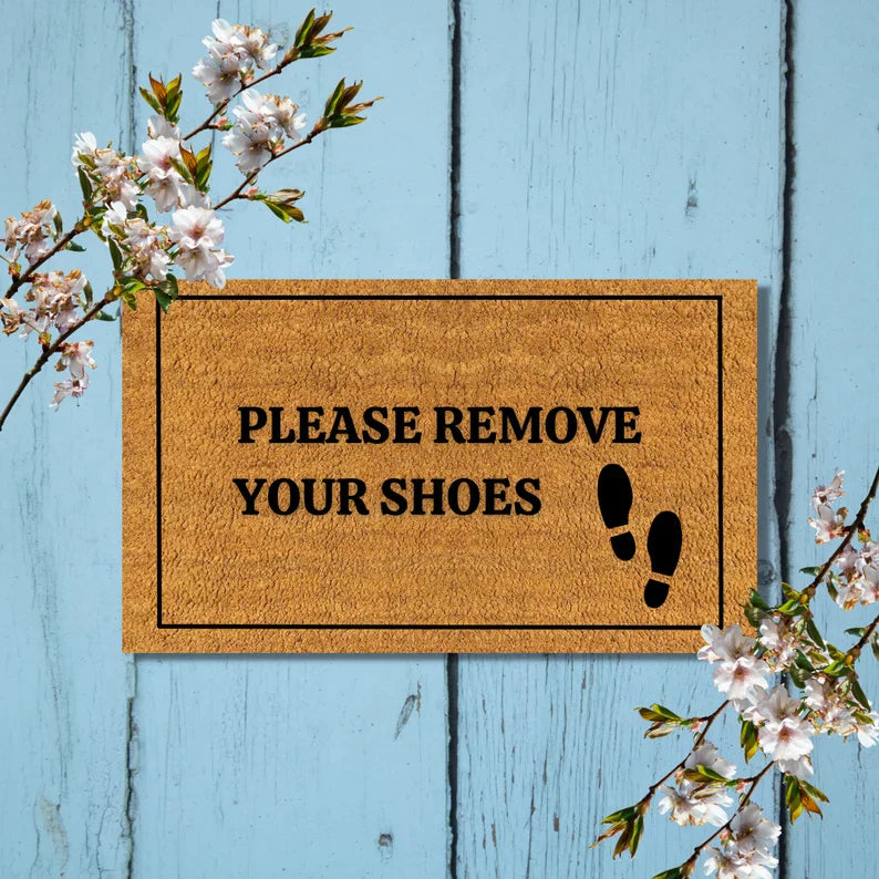Coir doormat with "PLEASE REMOVE YOUR SHOES" text and footprint graphic, displayed on blue wooden background with flowering branches