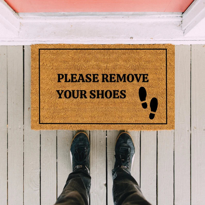 Tan coir doormat with black border and text reading "PLEASE REMOVE YOUR SHOES" with footprint graphic, positioned at entryway of white wooden door with pink ceiling trim.