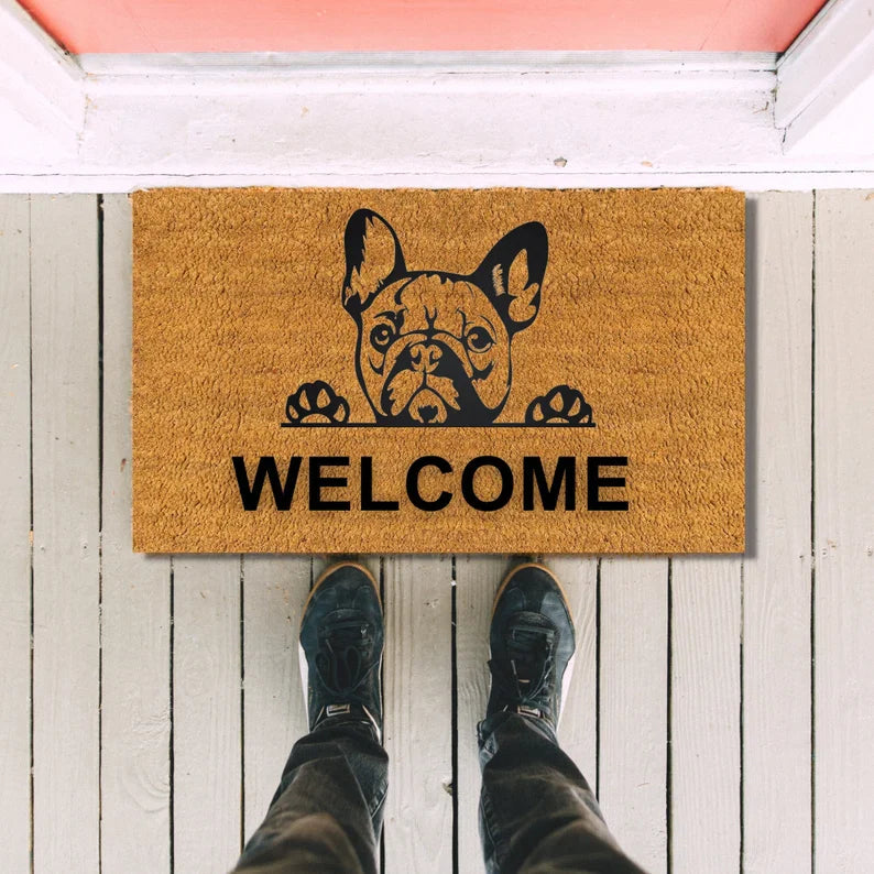 Tan coir doormat with black French Bulldog illustration and paws above the word "WELCOME," positioned on white wooden porch boards with person's feet in boots visible.