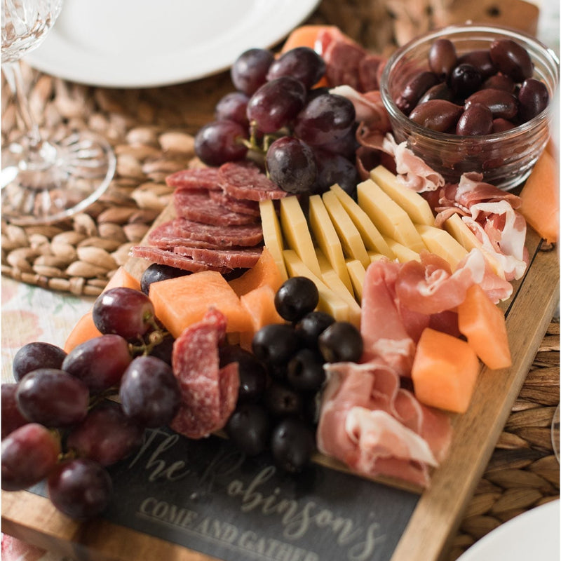 Wooden serving board with slate inlay styled with charcuterie, including grapes, cured meats, cheese, olives, and nuts arranged on surface