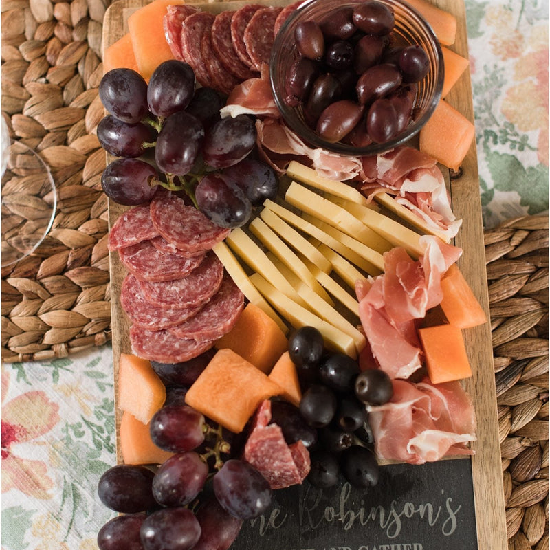 Wooden serving board with slate inlay engraved with sample text "The Robinsons Come and Gather," styled with charcuterie, grapes, cheese, cured meats, and olives
