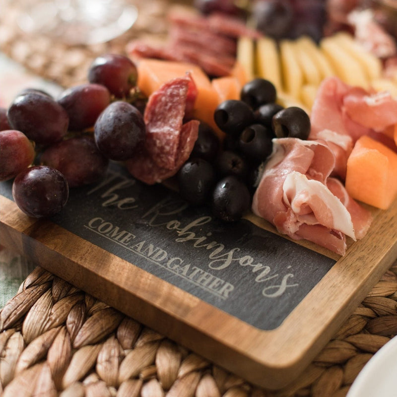 Slate and acacia wood serving board with sample engraving "The Johnsons' Come and Gather" styled with charcuterie, cured meats, olives, and cheese