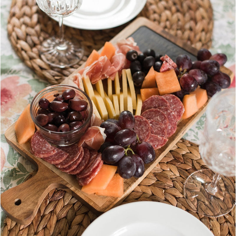 Wooden serving board with slate inlay arranged on woven placemats alongside charcuterie, grapes, cheese, olives, and glassware in a table setting.