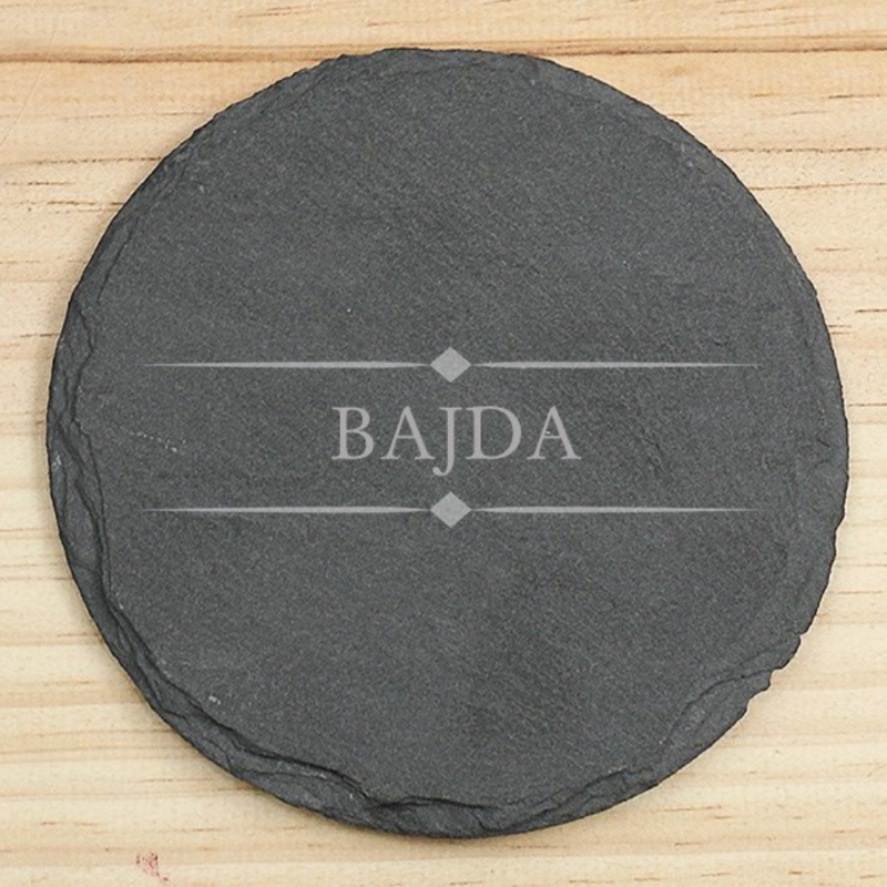 Round black slate coaster engraved with sample name "BAJDA" and decorative diamond flourishes, displayed on light wood surface