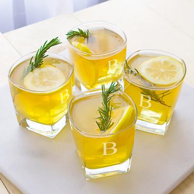 Four whiskey glasses filled with golden liquid and ice, each engraved with a sample letter "B", garnished with rosemary sprigs and lemon slices on a white table.