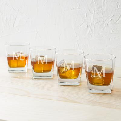 Four personalized whiskey glasses arranged in a row, each engraved with a sample monogram initial, filled with amber whiskey and ice on a light wood surface