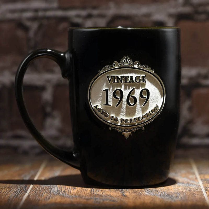 Black ceramic mug with ornate brass-colored oval medallion engraved with "Vintage 1969 Aged to Perfection," displayed on wooden surface with brick background.